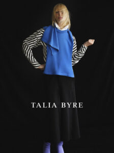 A person stands against a dark backdrop wearing a bright blue sleeveless top over a black and white striped shirt. They have long, light-colored hair partially covering their face. The outfit includes a long black skirt, complemented by light purple footwear. The scene conveys a sense of modern, avant-garde fashion, striking in its bold color contrasts.