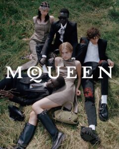 A group of five fashionably dressed adults lounge on dry grass in a relaxed, intertwined arrangement. Tailored jackets, structured dresses, polished shoes, and statement accessories create a refined yet undone mood. Their poses feel intimate and nonchalant, gazes drifting away from one another. Earthy tones meet sharp tailoring, suggesting modern aristocracy, quiet rebellion, cinematic calm, texture, and restrained drama.