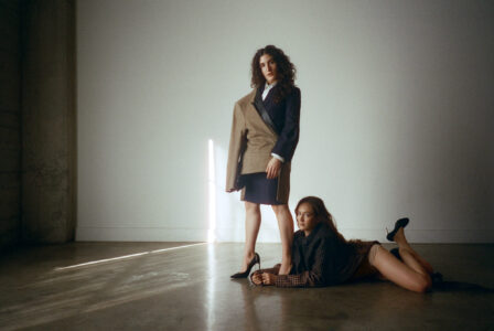 Two individuals are depicted in a minimalist setting, standing and reclining on the floor. One wears a mixed-fabric ensemble, holding one leg elevated. The other lies with legs crossed, dressed in a checkered jacket and beige shorts. Soft light casts shadows on the plain wall and floor, contributing to a serene and artistic ambiance.