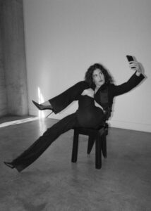 A person dressed in a dark outfit sits casually on a stool, with one leg extended and crossed over the other. They are holding a phone at arm’s length, appearing to take a selfie. The background is simple, with smooth walls meeting a concrete floor. The overall scene exudes a sense of relaxed modernity and confidence.