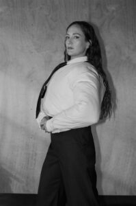 A grayscale representation features a person with long, flowing hair standing confidently against a wooden backdrop. The individual is dressed in a white shirt and dark pants, coupled with a tie. They maintain an assertive posture, with hands resting on their waist, exuding strength and poise in a composition that highlights simplicity and elegance.