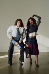 Two individuals strike dramatic poses indoors, one wearing an oversized suit with a loosened tie, holding a colorful object. The other wears a layered outfit, featuring a jacket over a blouse and a patterned skirt. Both are in high heels, standing on a polished floor with shadows cast from an unseen light source, evoking a theatrical atmosphere.