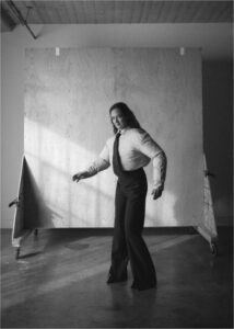 A person stands on a concrete floor in a room with a wooden wall or panel behind them. The lighting creates dynamic shadows and highlights. Wearing a light shirt, tie, and dark flared pants, the individual poses with slightly bent arms, exuding a confident expression. The ceiling has a textured design with visible industrial elements.