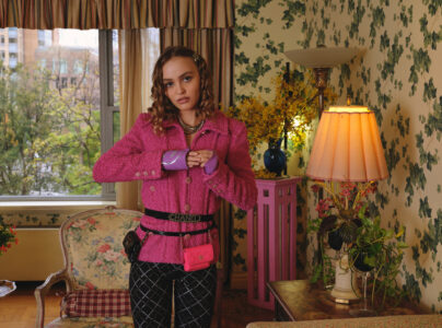 A young woman in a vibrant room stands confidently, wearing a textured pink jacket and plaid pants. The room features floral wallpaper, a large window with trees outside, elegant furniture, and a warmly lit lamp. Her hair has soft curls, and she accessorizes with purple gloves and a bright pink belt bag, creating a stylish and eclectic look.