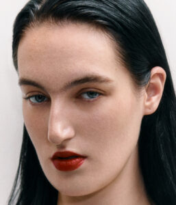 A person with sleek, dark hair and fair skin gazes intently. Their sharp features include well-defined eyebrows and striking, light-colored eyes. Vibrant red lips add a bold touch of color. The overall look is refined and elegant, emphasizing natural beauty and a confident, captivating presence. The composition is simple yet powerful, focusing on facial expression.