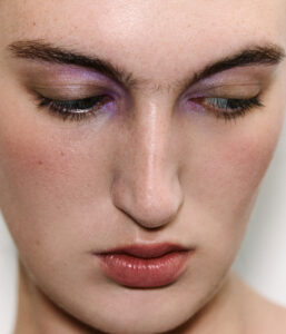 A close-up of a face reveals closed eyes adorned with shimmering purple eyeshadow, creating a soft, elegant look. The skin appears smooth with a natural complexion and subtle, warm undertones. The lips are gently pursed, showcasing a muted shade of natural pink. The overall expression conveys a serene and contemplative mood, enhanced by minimal additional makeup.