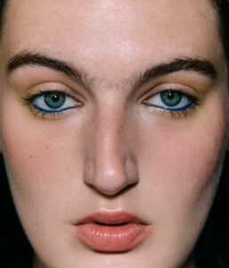 A close-up showcases a person's face, highlighting their clear blue eyes framed by bold, dark eyebrows. The smooth complexion reveals subtle freckles and a prominent nose. Their lips are slightly parted and have a natural, glossy appearance. Soft lighting accentuates the facial features, creating a clean and polished look with understated makeup and subtle warmth.