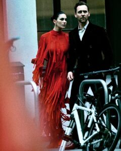Two stylish adults stride through an urban street, moving with purpose. One wears a dramatic red, fringed, body skimming outfit that ripples with motion, hair sleekly pulled back. The other is dressed in a dark tailored coat over a crisp shirt. Their expressions are focused and alert, bicycles framing the foreground, lending grit, contrast, tension, cinematic mood, rhythm, modern elegance.
