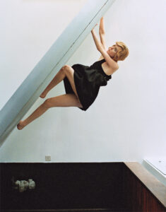A person in a black dress is suspended almost horizontally while gripping a slanted metallic structure. Their legs are positioned diagonally, bracing against the structure. The setting showcases a minimalist interior with white walls and a recessed wooden area below. The scene conveys a sense of balance, poise, and unique interaction with modern architecture.