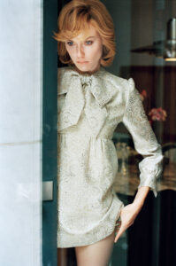 A person with short, light hair leans against a door in a contemplative pose. They're wearing an intricately patterned, light-colored dress featuring a large bow at the collar. The setting has a warm, indoor ambiance with soft lighting illuminating the figure. The background includes blurred objects, adding depth to the composition, enhancing the overall serene and thoughtful mood.