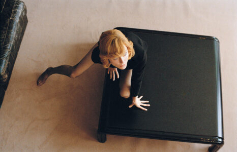A person with auburn hair, wearing a black dress, strikes a crawling pose on a sleek, dark, square table. One hand and one knee rest on the table, while the other hand supports on the floor. The setting features a beige carpeted floor and a dark, textured sofa to the side, creating a modern, stylish ambiance.