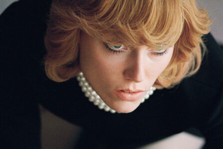 A close-up portrays a person with strawberry blonde hair and bangs, showcasing delicate facial features and a pensive expression. The individual wears a black garment accented with a strand of large white pearls. The lighting emphasizes their contemplative gaze and soft skin tone, creating an intimate and reflective mood. The composition is both stylish and emotive.