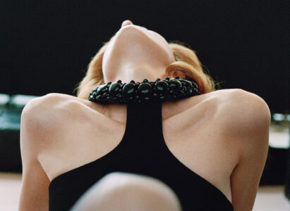 A person with fair skin is captured from below, leaning back with their neck extended. They are wearing a black, sleeveless garment. A bold, chunky, dark beaded necklace adorns their neck, creating a striking contrast. The background appears soft and blurred, emphasizing the subject's pose and accessories. Their light hair falls behind, enhancing the overall elegance.