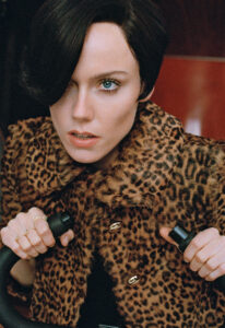 A person with intense blue eyes and dark hair gazes forward, exuding confidence and mystery. Dressed in a leopard print coat with golden buttons, they grip the handles of an object. The fur-patterned fabric adds a touch of luxury, while the bold rings on their fingers highlight a sense of elegance and style.