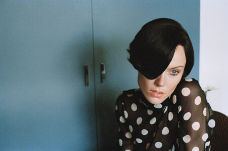 A person with striking features and blue eyes gazes intently. They have dark hair styled to cover one eye, creating a mysterious look. The polka-dotted, semi-transparent top adds a retro flair. The background features a muted blue cabinet, enhancing the scene's subtle elegance. The lighting casts soft shadows, highlighting the expressive expression and stylish attire.