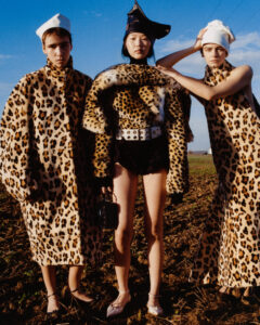Three individuals stand on a grassy field wearing leopard print outfits. Two have long coats, one wears a short jacket with furry trimming. They sport various hats, including a white sailor-style cap and a black leather piece. The shoes are elegant with delicate straps. The bright blue sky creates a striking contrast, adding an artistic flair.