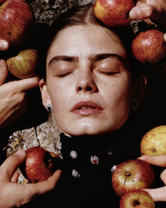 A serene face with closed eyes is surrounded by several hands gently holding apples. The setting is earthy with a textured stone background. The person wears a black outfit adorned with sparkling embellishments. The composition is calm, with a warm and natural color palette, evoking a sense of tranquility and connection with nature.