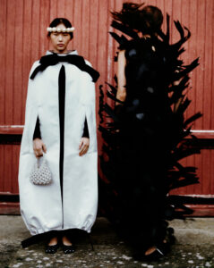 Two individuals stand against a textured, red backdrop. One wears a voluminous white garment with black accents, holding a patterned bag and donning a pearl headpiece. The other is clad in a dramatic black outfit adorned with protruding feather-like shapes. Both stand on a stone surface, exuding avant-garde fashion flair and contrasting light and dark tones.