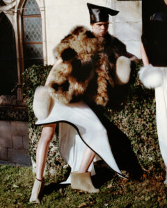 A stylish individual poses with confidence, draped in a luxurious fur garment that contrasts with their sleek white outfit and ankle boots. They wear a distinctive black cap, standing against a backdrop of ivy-covered stone walls and medieval-style windows. The scene is bathed in natural light, casting gentle shadows that enhance the texture of the ensemble.