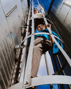 Two individuals dressed in stylish attire are climbing a metal ladder attached to a modern industrial structure. The scene is captured from a low angle, emphasizing the towering height and capturing a vivid blue sky. One person wears a white top, the other a blue garment and plaid pants. The setting conveys an adventurous and dynamic energy.