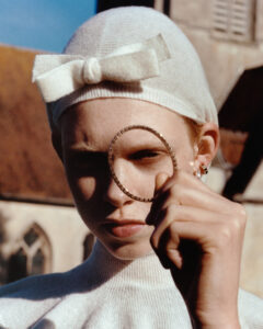 A person holds a circular object up to one eye, squinting slightly. They wear a white cap with a bow and a matching turtleneck sweater. The background features a blurry architectural structure with pointed windows and a blue sky, suggesting an outdoor setting. The overall vibe is vintage and artistic, highlighting the individual's unique style and expression.