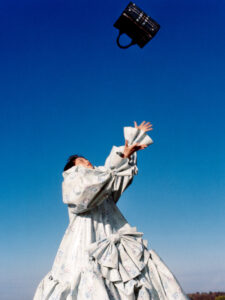 A person stands against a vibrant blue sky, wearing a voluminous, floral-patterned dress with large, dramatic bows. They gaze upwards with outstretched arms reaching toward a dark handbag floating above. The scene has an open, airy feeling, with the expansive sky creating a sense of freedom and movement, contrasting with the intricate detailing of the dress.