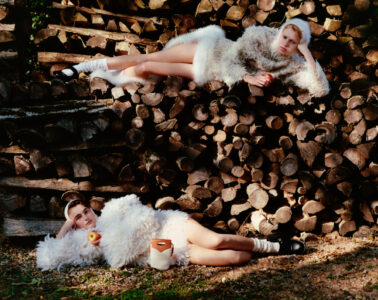 Two individuals are reclining on a large stack of chopped logs, surrounded by a natural outdoor setting. They wear fluffy white garments and black shoes with white socks. Each holds an apple, contributing to a whimsical, rustic aesthetic. The scene includes light and shadow play, emphasizing the textured wood and clothing, creating a serene and artistic composition.
