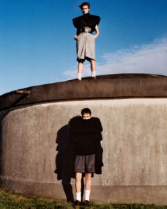 Two individuals are positioned uniquely against a concrete structure, one standing atop the curved surface, the other leaning below. Both wear avant-garde clothing, mixing oversized black tops with tailored shorts and shoes. Their shadows extend on the textured wall, contrasting against the blue sky. The scene blends natural and architectural elements, suggesting a blend of modernity and contemplation.