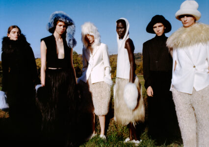 A group of six people dressed in avant-garde fashion stand in a natural setting. They wear various outfits with fluffy, furry textures, bold colors, and oversized accessories like hats and collars. The scene is bright with a clear blue sky in the background, creating a striking contrast against the greenery of the outdoors.