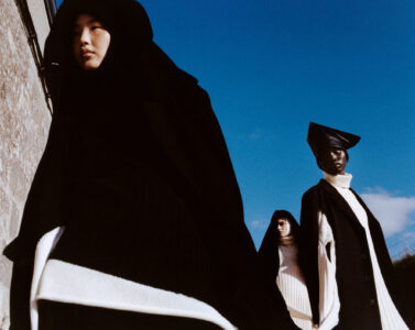 Three individuals stand against a vivid blue sky. They are dressed in striking black and white ensembles that feature dramatic silhouettes. The first person, cloaked in black, gazes forward. The second wears a unique headpiece and a layered outfit with a high collar. The third, partially obscured, also embraces a monochromatic aesthetic. A building with a stone texture forms the background.