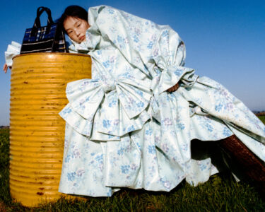 A person in an elaborate, floral-patterned outfit leans against a yellow, textured barrel amidst a grassy field under a clear blue sky. The outfit features large bows and voluminous sleeves. A striped bag rests atop the barrel. The scene combines contrasting colors and textures, creating a visually striking composition against the natural backdrop.