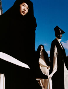 Three individuals stand under a bright blue sky, wearing dramatic black cloaks and white garments with bold, contrasting designs. Each person displays unique headwear and attire that blend modern and traditional styles. The angular and flowing fabrics create a striking visual effect, emphasizing texture and form. Their expressions are serene, adding a sense of elegance and mystery.