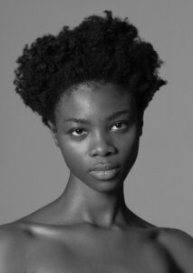 A black-and-white portrait features an individual with natural curly hair styled into a voluminous shape. The expression is calm and direct, with eyes softly engaging the viewer. The lighting highlights the smooth texture of the skin, creating subtle contrasts and emphasizing facial features. The composition is simple, focusing entirely on the subject against a plain backdrop.