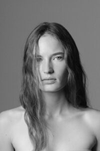 A person gazes directly ahead, exhibiting a composed expression. Their long hair cascades gently over one shoulder, partially covering the face. The setting is minimalistic, with a neutral background emphasizing the serene and contemplative atmosphere. The monochrome tones enhance the figure's natural features and exude a timeless, classic elegance.