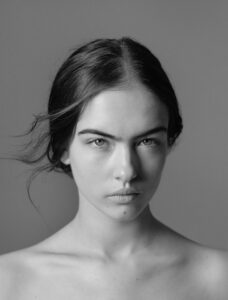 A monochrome portrait features a young person with a serious expression. Their hair is dark and parted in the middle, with a few strands falling loosely. The gaze is direct and intense, capturing attention. The background is plain and unobtrusive, emphasizing the subject's facial features and composed demeanor. The lighting creates gentle shadows, adding depth.