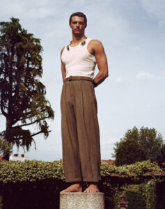 A person stands barefoot on a pedestal against a backdrop of greenery and a partly cloudy sky. Wearing a white sleeveless top with colorful details at the neck and loose brown trousers, the individual exudes confidence. The scene is peaceful, with tall trees and manicured shrubs enhancing the serene outdoor setting, creating a sense of harmony with nature.