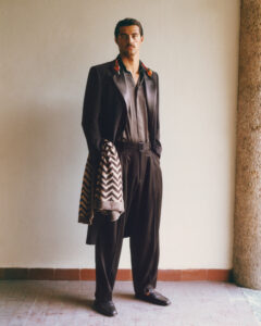 A stylish individual stands confidently in a minimalist setting. They are dressed in a smart, dark ensemble featuring a sleek jacket, button-up shirt, and belted trousers. A patterned piece of fabric is draped over an arm, complementing the outfit. The floor consists of rustic tiles, and a rough-textured column is positioned on one side.