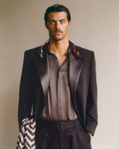 A man stands confidently against a neutral backdrop, wearing a dark suit with satin lapels over a silky shirt. A colorful cloth is draped around his neck. He sports a mustache and has his hair slicked back. In his right hand, he holds a patterned fabric, while his left hand is casually in his pocket.