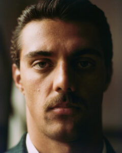 The portrait features a person with a focused expression, neatly groomed brown hair, and a prominent mustache. The lighting highlights strong facial features and creates soft shadows, lending a moody atmosphere. The subject wears a formal outfit, visible through part of a crisp white collar. The background is a blurred, warm tone, emphasizing the subject's presence.