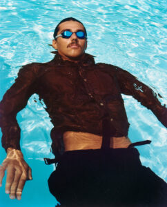 A man floats effortlessly on his back in a clear blue pool, wearing sunglasses and a dark shirt. Sunlight dances across the water, creating shimmering patterns on his clothing. His hair is slicked back, and a sense of calm pervades the atmosphere. The scene captures a moment of relaxation and serenity, enhanced by the vibrant aquatic surroundings.
