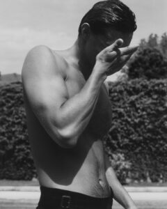 A person stands shirtless, their body glistening as it catches the light. They're captured mid-action, with one hand raised to their face. The background shows a blurred landscape of foliage and sky, suggesting an outdoor setting. The overall tone is monochromatic, emphasizing contrasts in light and shadow, creating a sense of depth and focus on the subject.