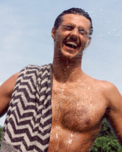 A shirtless individual stands joyfully under running water, smiling broadly with eyes closed. Water droplets glisten on their skin as sunlight illuminates the scene. A towel with a zigzag pattern is draped over their shoulder. The sky above is clear and blue, while trees in the background hint at a sunny outdoor setting, enhancing the refreshing atmosphere.