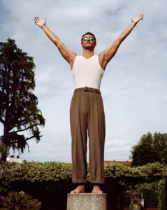 A person stands barefoot on a stone pedestal, arms outstretched upward against a cloudy sky. They wear sunglasses, a white tank top, and high-waisted brown trousers. The background features lush greenery, including tall trees and shrubs, creating a serene, natural setting that enhances the posture's expression of freedom and openness.