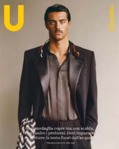 A man with a serious expression stands against a neutral background. He wears a dark blazer over a silky shirt, and a patterned scarf is draped across one shoulder. Swim goggles hang around his neck, adding a touch of color. His hair is slicked back, and he sports a neat moustache, exuding a confident and stylish aura.