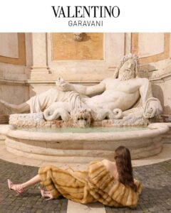 A grand stone fountain featuring a reclining classical figure dominates a sunlit square. In the foreground, a woman draped in a voluminous golden garment reclines on cobblestones, her flowing hair echoing the sculpture’s elegance. Soft neutral tones and monumental architecture create a dialogue between fashion, history, and theatre, blending contemporary glamour with the timeless grandeur of the surrounding setting.