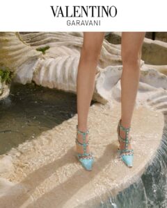 A person stands on the edge of a stone surface, surrounded by gently flowing water. Their feet are adorned with elegant, pointed turquoise heels with straps and metal studs. The setting includes natural rock formations in a pale, textured hue, giving the scene an artistic and sophisticated feel, perfectly combining fashion with a natural backdrop.