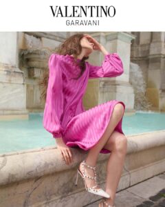 A person sits gracefully on the edge of a fountain, elegantly dressed in a vibrant pink long-sleeve dress that features a subtle striped pattern. The individual wears white high-heeled shoes adorned with studs. The background reveals intricate stone architecture and cascading water, creating a serene and stylish atmosphere. The setting exudes a sense of luxury and fashion.