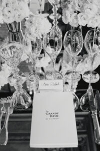 A formal dining setup features elegantly arranged glassware including wine glasses and a decorative goblet. The table is adorned with lush floral arrangements in the background, composed of soft, voluminous blooms. Cutlery is meticulously placed on either side, reflecting a sophisticated atmosphere. The monochrome tones add a timeless and classic feel, evoking a sense of refined elegance.