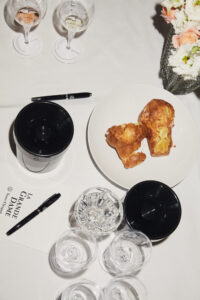 A white tablecloth hosts two glasses of wine, one rosé and one white, surrounded by several empty glasses. A black cup sits near a pair of ink pens. A white plate holds two fluffy pastries, while a vase brimming with white and peach flowers adds a soft touch to the setting.