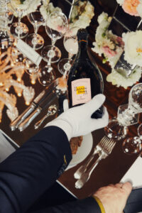 A well-dressed individual in formal attire holds a champagne bottle over an elegant table set with crystal glassware and silver cutlery. The setting is enhanced by delicate floral arrangements and warm lighting, creating a luxurious atmosphere. The composition captures a moment of sophistication and celebration, with the reflections on the table adding to the opulent ambiance.